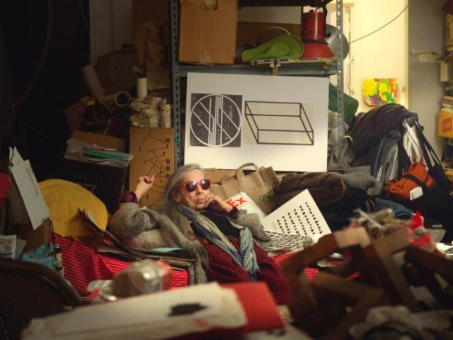 A woman sits amidst a cluttered room