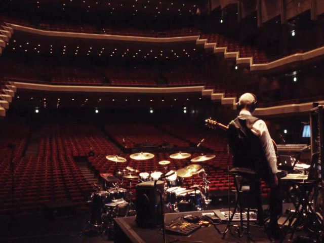 Robert Fripp stands on an empty stage