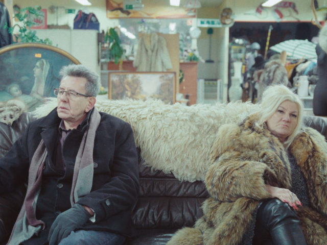 an older man and woman sitting on a sofa in a pawnshop and are smoking. The woman on the right has white hair and is wearing a fluffy fur coat, the man on the left wears glasses and a leather jacket.